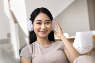 Kulaklıklı mutlu Asyalı alet kullanıcısı kız portresi. Kablosuz kulaklıklara dokunan, dişlek gülümsemeyle kameraya bakan genç bir kadın. Video çağrısı ekranı
