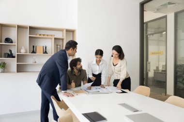 Çeşitli iş arkadaşları proje üzerinde birlikte çalışıyor, kağıt raporlarıyla masada buluşuyor, proje istatistiklerini analiz ediyor, dökümanları diyagramlarla kontrol ediyor, satış verileri üzerinde konuşuyor.