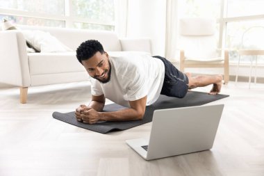 Neşeli güçlü atletik Afrikalı adam yoga minderinde statik kalas tutuyor, ekrana bakıyor, gülümsüyor, ders seyrediyor evde spor yapmak için internet iletişimini kullanıyor.