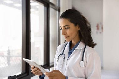 Ciddi genç doktor kadın iş için tablet kullanıyor, hastayla online bağlantı kuruyor, klinik başvurusunda elektronik randevu çizelgesini kontrol ediyor, dijital cihazla pencerenin yanında duruyor.