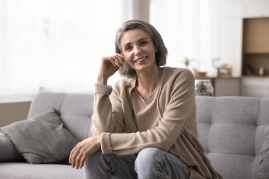 Neşeli olgun bir kadın kanepede dinleniyor, kameraya bakıyor çekici bir görüntüsü var, kusursuz beyaz bir gülümsemesi var, evde fotoğraf için poz veriyor, mutlu ve memnun görünüyor, huzurlu ve kendinden emin görünüyor, hafta sonunun tadını çıkarıyor.
