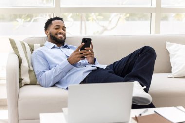 Keyifli, rahat Afrikalı serbest çalışan adam ofis koltuğunda dinleniyor, dinleniyor, keyfine bakıyor, akıllı telefondan online iletişim kuruyor, iş yerindeki laptopuyla cep telefonunda daktilo yazıyor.