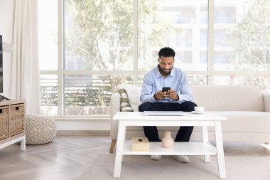 Yakışıklı Afrikalı serbest çalışan iş adamı akıllı telefonuyla yazı yazıyor, rahat bir evde kanepede oturuyor, çevrimiçi iletişimden hoşlanıyor, sohbet ediyor, mesaj yazıyor, internet teknolojisi kullanıyor.