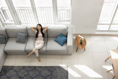 Rahatlamış, genç, Asyalı bir kadın. Gözleri kapalı bir şekilde evdeki büyük rahat koltukta dinleniyor. Rahatlamanın, boş zamanların, açık dirseklerle temiz hava solumanın tadını çıkarıyor. Üst görüntü görüntüsü