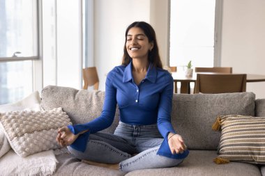 Huzurlu genç Hintli kadın meditasyon yapıyor, evde nefes alıyor, kanepede meditasyon yapıyor, parmaklarını jana mudra 'da tutuyor, enerji alıyor, uyum sağlıyor, konsantrasyon eğitimi alıyor.