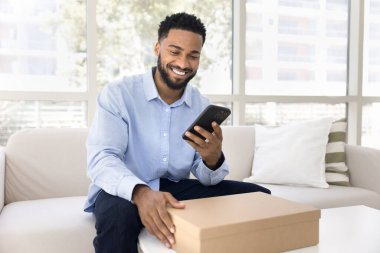 Lojistik karton kutunun üzerinde cep telefonuyla yazı yazan, teslimat servisinden paket alan akıllı telefon kullanan, gülümseyen ve gülen genç Afrikalı genç bir müşteri.