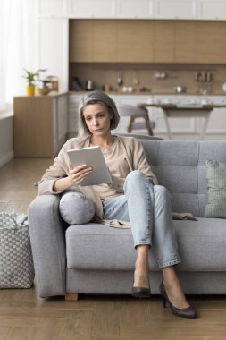 Odaklanmış yaşlı kadın haftasonunu dijital tablet cihazıyla oturma odasında oturarak, tatil planları yaparak, bilet ya da eşya alarak, internette gezinerek, e-postaları evde boş vakitlerle kontrol ederek geçiriyor.