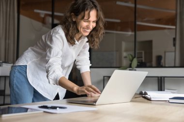 Neşeli genç iş kadını laptopta iş iletişiminden zevk alıyor, iş yerinde bilgisayarla duruyor, daktilo ediyor, gülüyor, video görüşmesinde konuşuyor.