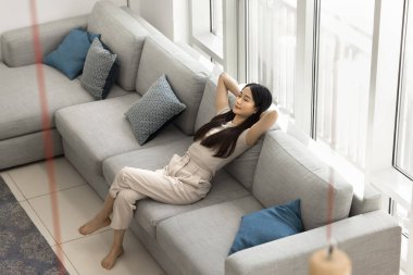 Calm beautiful Asian adult girl enjoying relaxation at cozy home, resting on comfortable spacious grey couch with hands on nap, taking deep breath of fresh air with closed eyes. High angle shot clipart