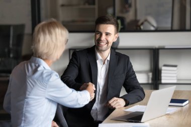 Resmi gömlekli neşeli genç bir iş adamı olgun bir iş kadınıyla el sıkışıyor çalışma masasında oturuyor, gülümsüyor, danıştıktan sonra müşteriyle tokalaşıyor.
