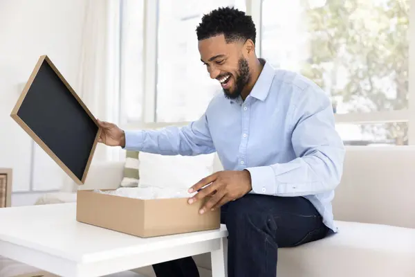 エキサイティングな幸せな若い黒人の消費者は自宅で段ボール箱を開け 荷を解くことはオンラインショップからの購入を待って 論理的な小包を得 驚きで笑います — ストック写真
