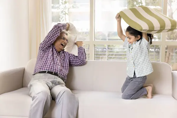 Excited joyful Indian grandpa and little granddaughter girl fighting with pillows on home couch, laughing, playing active games, enjoying battle, leisure, playtime. Granddad watching grandkid