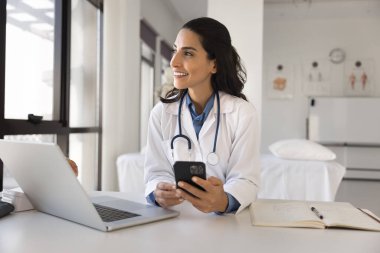 Mutlu genç doktor kadın akıllı telefonda online tıbbi uygulama kullanıyor, iş yerinde cep telefonu tutuyor, internette danışmanlık yapıyor, başka yere bakıyor, düşünüyor, modern teknolojiden zevk alıyor.