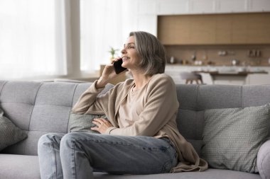 Gülümseyen, orta yaşlı, çekici bir kadın oturma odasındaki kanepede dinleniyor, başka tarafa bakıyor, cep telefonunu tutuyor, arıyor, yakınlarıyla hoş bir uzaktan konuşma yapıyor, bilgi paylaşıyor, iletişimin tadını çıkarıyor.