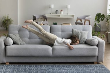 Sevimli küçük kız modern evde kaygısızca eğleniyor, oturma odasındaki rahat kanepeye atlıyor, rahat mobilyalarda eğleniyor, mutlu görünüyor, hafta sonları zevk alıyor. Yorgunluk giderme, rahatlama.
