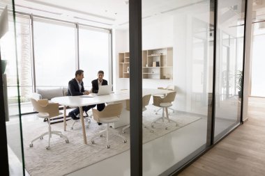 İki iş adamı online proje üzerinde birlikte çalışıyorlar, modern toplantı odasında dizüstü bilgisayarla konuşuyorlar, beyaz ofis içinde dizüstü bilgisayar kullanıyorlar, işbirliği yapıyorlar, ağ kuruyorlar.