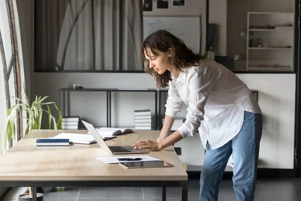 stock image Focused Caucasian young entrepreneur woman in casual clothes working on project in office alone, typing on laptop, standing at workplace, using modern Internet technology for job tasks