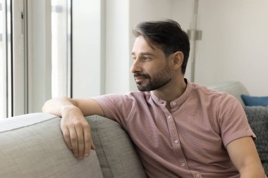 Olumlu düşünceli, rahat orta yaşlı bir adam rahat koltukta oturuyor, pencereye bakıyor, düşünüyor, hayal kuruyor, evde dinleniyor, sessizleşiyor, rahatlıyor, rahatlıyor, gülümsüyor.