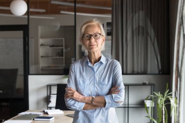 Zarif gözlüklü, kendine güvenen, kolları bağlı, modern ofis işyerinde duran, kameraya bakan, kendine güvenen, kıdemli bir iş kadını. Olgun yönetici, profesyonel portre için yönetmen.