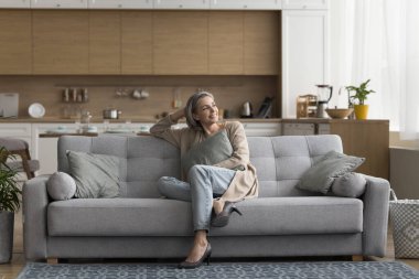 Olumlu gülümseyen olgun kadın oturma odasındaki rahat koltukta dinleniyor, başka yere bakıyor, evde tek başına vakit geçiriyor, hafta sonunun keyfini çıkarıyor, rahat bir hayat sürüyor, izin günlerinde veya kiralık bir dairede hayal kuruyor.