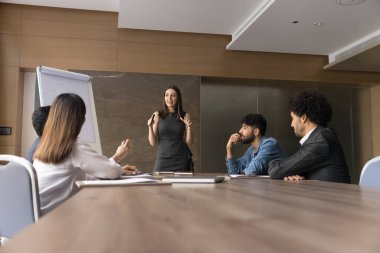 Heyecanlı proje lideri kadın, çok ırklı takımlara başlangıç fikirleri sunuyor, toplantı masasında duruyor, yaratıcı planı tartışıyor, oturmakta olan meslektaşları ve çalışanları ile strateji tartışıyor.