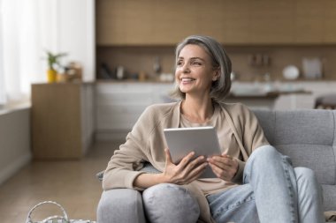 Mutlu, orta yaşlı kadın dijital tablet tutuyor, rahat bir koltukta oturuyor, başka yere bakıyor, sosyal medyada sohbet ediyor, online iletişimden hoşlanıyor, düşünüyor, planlıyor, internette hobi için uygulama kullanıyor.