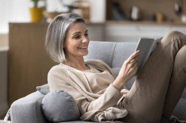 Carefree happy middle-aged woman spend time at home, read interesting book, lies on comfortable couch, enjoy leisure weekend with favorite novel, booklover rest on sofa in living room, entertainment clipart