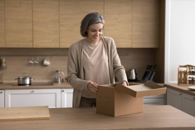Gülümseyen kadın mutfak masasında duruyor açık paket kutusu, evdeki internet mağaza siparişini açıyor, hızlı teslimat hizmetlerinden memnun, nitelikli mallardan memnun, internetten alışveriş yapıyor.