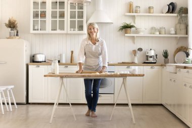Tatlı gülümsemesi için ev yapımı hamuru pişiren olgun kadın portresi kameraya bakıyor, sıcak, modern evcil mutfakta yemek hazırlıyor. Ev hanımı işleri, aşçılık, doğal ürünler kullanan aile tarifi.