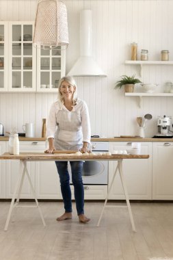 Güzel orta yaşlı bir kadın modern hafif mutfakta tek başına önlük giyer çörekler için hamur ezer, gülümser, hafta sonları kameraya poz verir, yemek pişirirdi. Ev işi, ev yapımı pişirme.