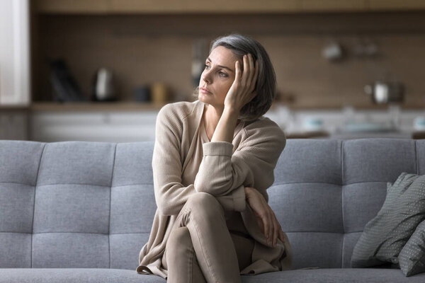 Attractive silent middle-aged woman relaxing seated on couch in living room, staring out window, ponders, missing, deep in thoughts, looks pensive and thoughtful, spend boring weekend alone at home