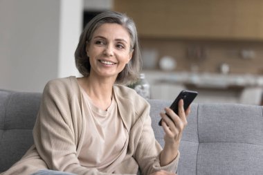 Neşeli, olgun bir kadın oturma odasındaki rahat koltukta dinleniyor, akıllı telefonu tutuyor, başka yere bakıyor, gülümsüyor, arkadaşını arıyor, mesajını paylaşıyor, iyi haberleri düşünüyor, yeni uygulama kullanıyor, interneti kullanıyor.