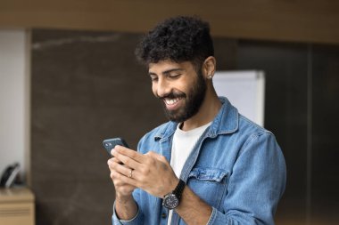 Mutlu genç Arap proje yöneticisi cihaz üzerinde online uygulama kullanıyor, gülümsüyor, gülüyor, cep telefonunda daktilo yazıyor, internet iletişiminden keyif alıyor, medya hizmetlerinden faydalanıyor.