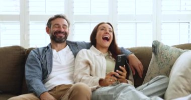 Sevimli Portekizli çift kanepelerinde modern akıllı telefonlarıyla rahatlıyor, komik videolara gülüyorlar, online içeriği izliyorlar, evde kaygısızca vakit geçiriyorlar, yeni cep telefonu uygulamaları kullanıyorlar.