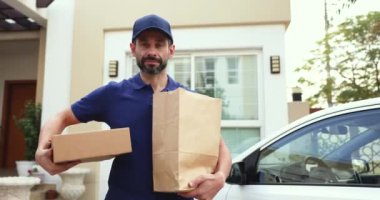 Mavi tişörtlü ve kep üniformalı teslimatçı elinde paket çantaları ve paket kutusuyla müşteri evinin yakınındaki sokakta poz veriyor, gülümsüyor, kameraya bakıyor, mükemmel nitelikli müşteri hizmetleri sunuyor.