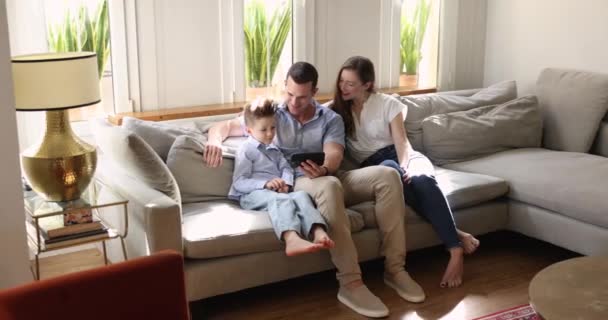 父母和8岁的小儿子笑着 用手机在沙发上放松 享受着新的娱乐应用 在舒适的公寓里度过了一个愉快的周末 使用现代小玩意的家庭休闲时间 免版税图库视频片段