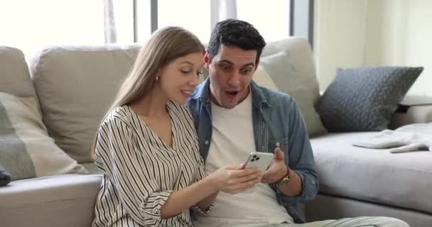 令人惊讶的千年西班牙裔妻子和丈夫坐在客厅里 拿着智能手机 在Sms上看到好消息 网上彩票中奖 银行按揭审批 年轻夫妇的新工作机会 — 图库视频影像