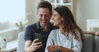 Mutlu İspanyol genç çift haftasonunu evde oturarak, oturma odasında cep telefonuyla oturuyor, yeni cep telefonu uygulamalarıyla eğleniyor, sosyal medyada komik videolara gülüyor, şakaya gülüyor.