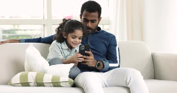 愛する若いインドの父親は かわいい息子とソファーに座ってインターネット上で自由な時間を過ごし 新しいモバイルアプリケーションを使用して一緒に楽しんで オンラインゲームをプレイし 仮想楽しいブログコンテンツを楽しんでいます — ストック動画