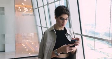 Cep telefonunda iş uygulamasını kullanan, mesaj yazan, akıllı telefondan internete bakan, gülümseyen, ofis penceresine bakan, odaklanmış genç Ortadoğulu profesyonel kadın.