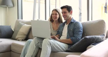 Genç İspanyol kadın ve erkek rahat bir oturma odasında dizüstü bilgisayarla oturuyor, İnternet üzerinden alışveriş yapıyor, online alışverişi tartışıyor, cihaz ekranına bakıyor, e-servis satın alıyor.