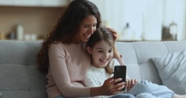 Genç mutlu kadın ve küçük kız komik videolara gülüyor, yeni mobil uygulamalar kullanıyor, akrabalarla video görüşmesinden zevk alıyor, modern uygulama ve akıllı telefonla evde zaman geçiriyor. Aile eğlencesi, teknoloji.