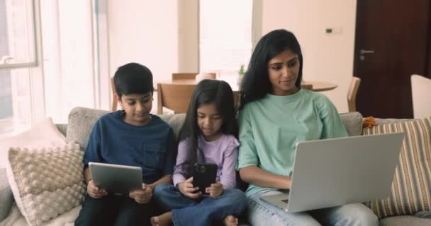 年轻妇女的小孩们坐在家里的沙发上 使用各种各样的电子设备 家人在网上消磨时间 使用平板电脑 笔记本电脑和手机进行娱乐和交流 小武器上瘾 — 图库视频影像