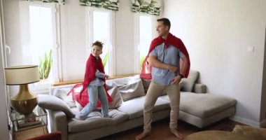 Kaygısız baba oğul dansı, evde birlikte oyun. Küçük çocuk ve baba kırmızı pelerin giyiyor süper kahramanlar gibi davranıyorlar, komik oyun zamanları geçiriyorlar, pop müziğe geçiyorlar, zıplıyorlar, oturma odasında oynuyorlar, uzun süre oynuyorlar.
