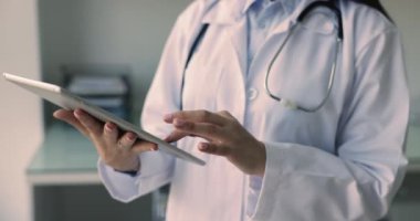 Dijital tablet tutan kadın pratisyen hekim, çevrimiçi çalışıyor, hastalar hakkındaki bilgileri gözden geçiriyor, tedavi planı yazıyor, rapor yazıyor, müşterilerle uzaktan terapi seansları yapıyor, teleoterapi uygulamaları kullanıyor.
