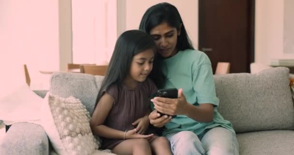 美丽的印度女人花时间和可爱的小女儿坐在沙发上 用现代智能手机 享受新的有趣的移动应用程序 教孩子如何使用小工具 打电话 在网上玩得开心 — 图库视频影像