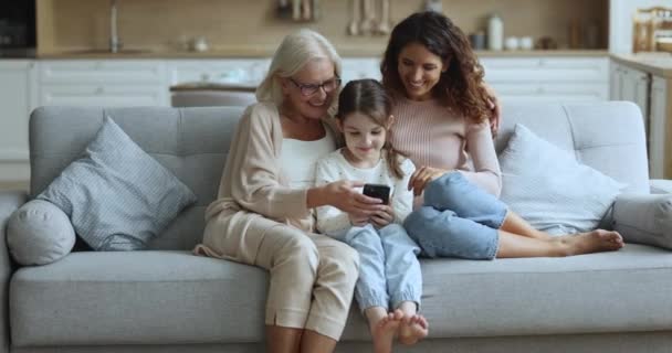 3世代の女性は 携帯電話付きの居心地の良いソファーで家でリラックスしたり 面白いオンラインコンテンツやビデオを見たり 現代のアプリケーションを使用したり 自宅で楽しい家族のレジャーを楽しんだりします — ストック動画