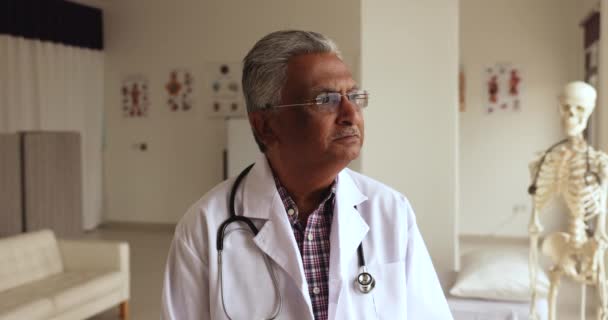 白いコートを着た真剣な思慮深い古いインドの医師は 目をそらして 医療専門職のキャリアを考え 幸せになり 笑顔になり クリニックオフィスに立っています — ストック動画