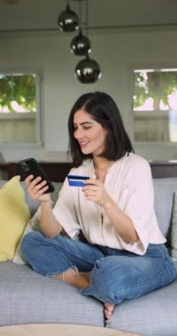 Latin bir kadın kart ve telefonla evdeki kanepeye oturup internetten alışveriş yapıyor. Güzel bir bayanın perakende ürün satın almasına, dijital abonelikleri yenilemesine ya da yayın hizmetleri platformlarına dikey bir bakış açısı. E-alışveriş