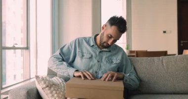 Genç Hispanik adam, elektronik ticaret müşterisi oturma odasındaki koltukta oturup paketleri açıyor ve internetten sipariş almaktan memnun oluyor. Elektronik hizmetler, ekspres nakliye